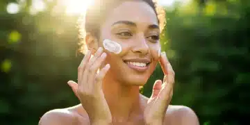 Mulher com pele radiante aplicando protetor solar no rosto sob o sol, representando cuidado e proteção diária para uma pele jovem.